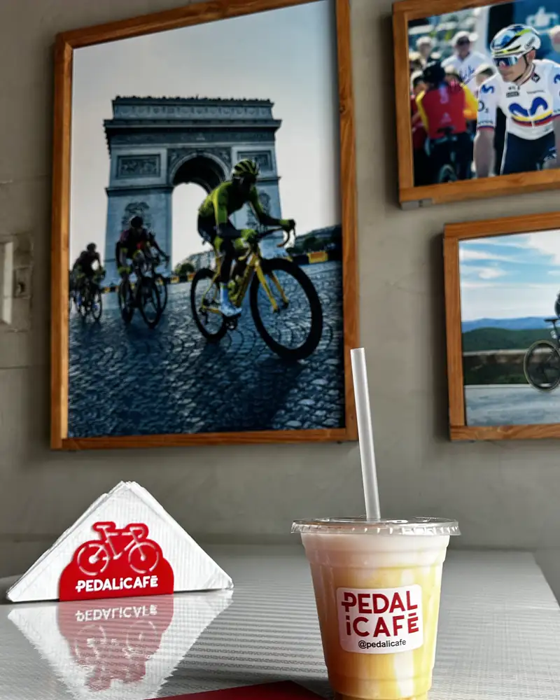 Bebida fría de PEDALiCAFÉ en una mesa; detrás, un cuadro de ciclistas del Tour de Francia frente al Arco del Triunfo