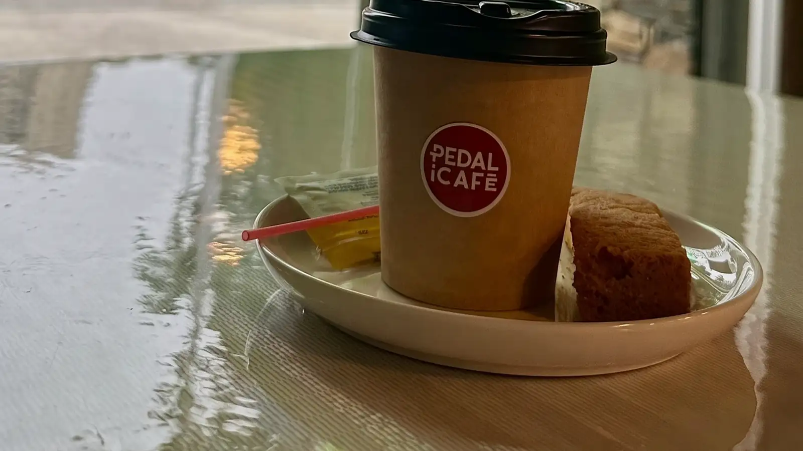 Café para llevar de PEDALiCAFÉ y un brownie sobre una mesa mojada junto a la ventana