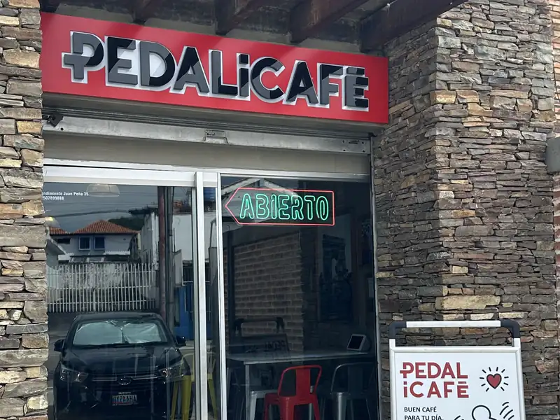 Fachada de PEDALiCAFÉ con el letrero rojo sobre la entrada de vidrio, la pared de piedra y la pizarra en la acera
